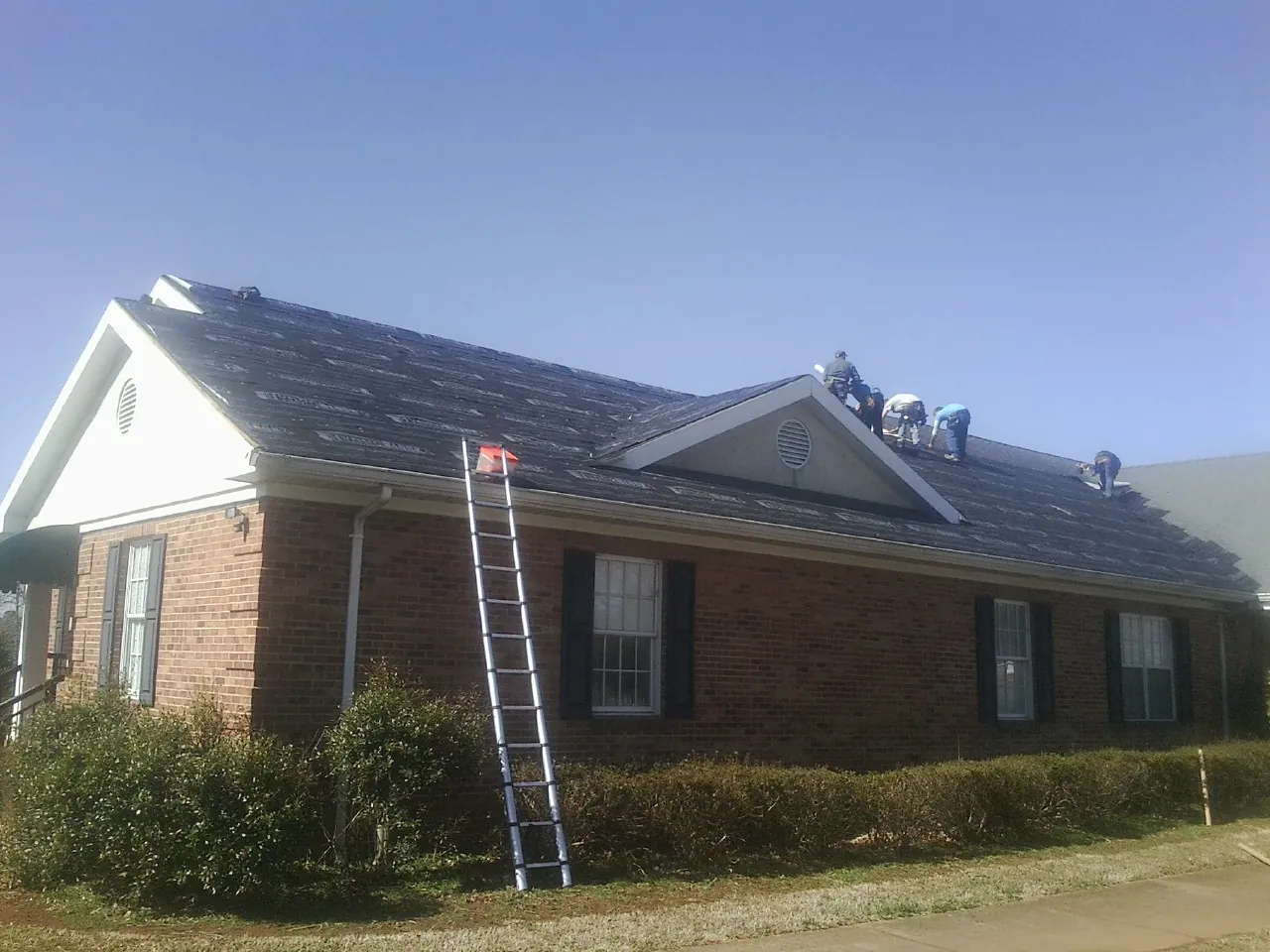 Crew tearing shingles off a brick ranch roof with a ladder set — Emergency Roof Repair work by our crew in Conyers