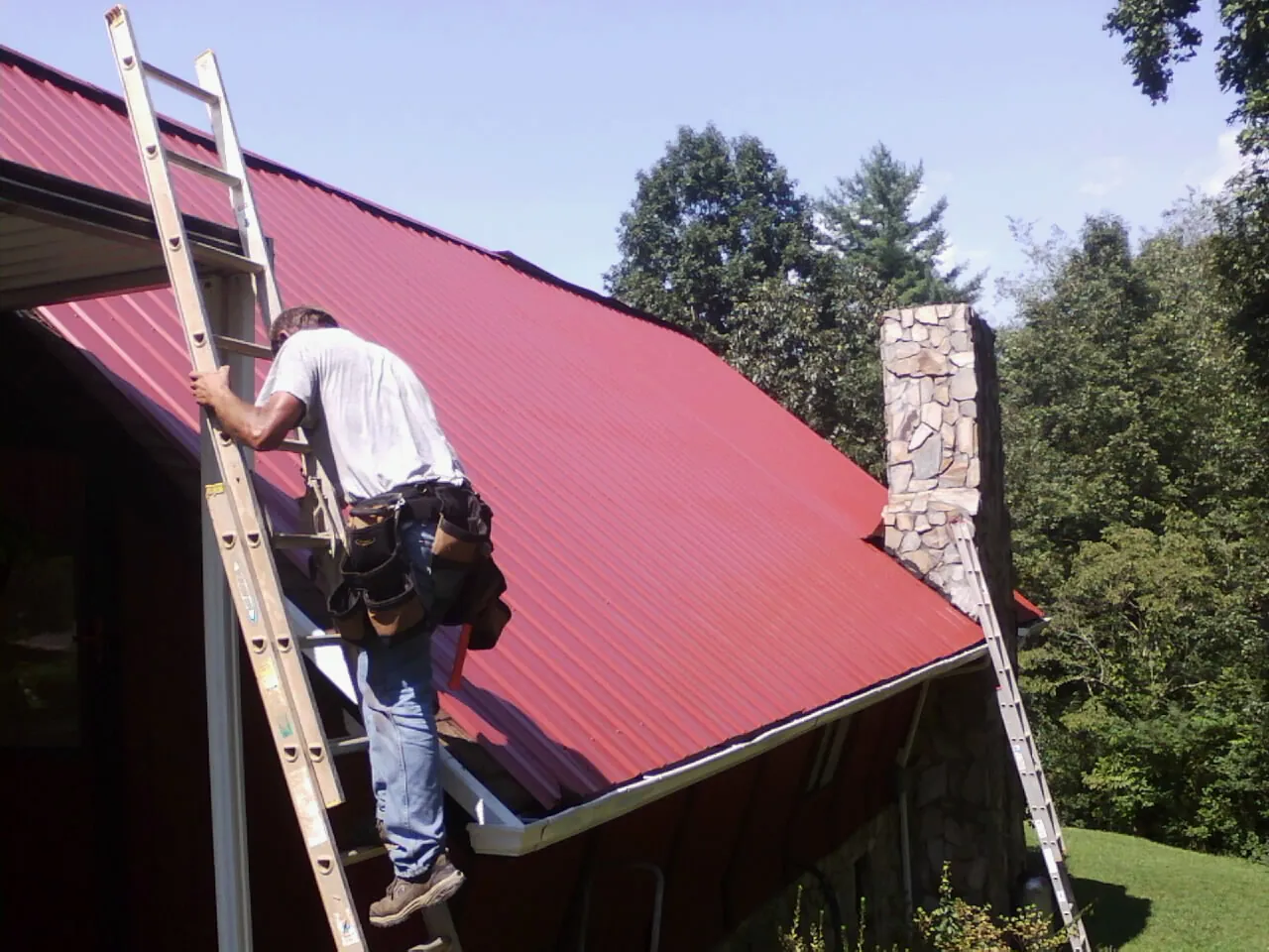 Roofer on a ladder beside a red metal roof and stone chimney — Soffit & Fascia Repair by Roofing Brothers in Conyers